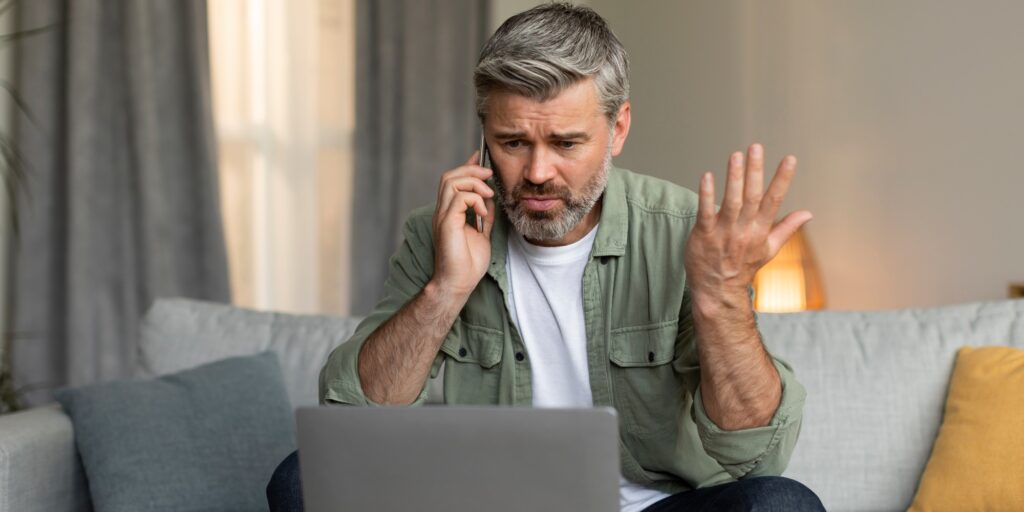 Sad angry elderly caucasian male calls by phone, yells and looks at laptop, gesticulates, has