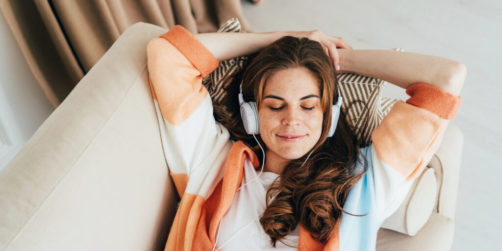 Young relaxed woman is laying down on sofa and listening to a music with headphones on her ears.